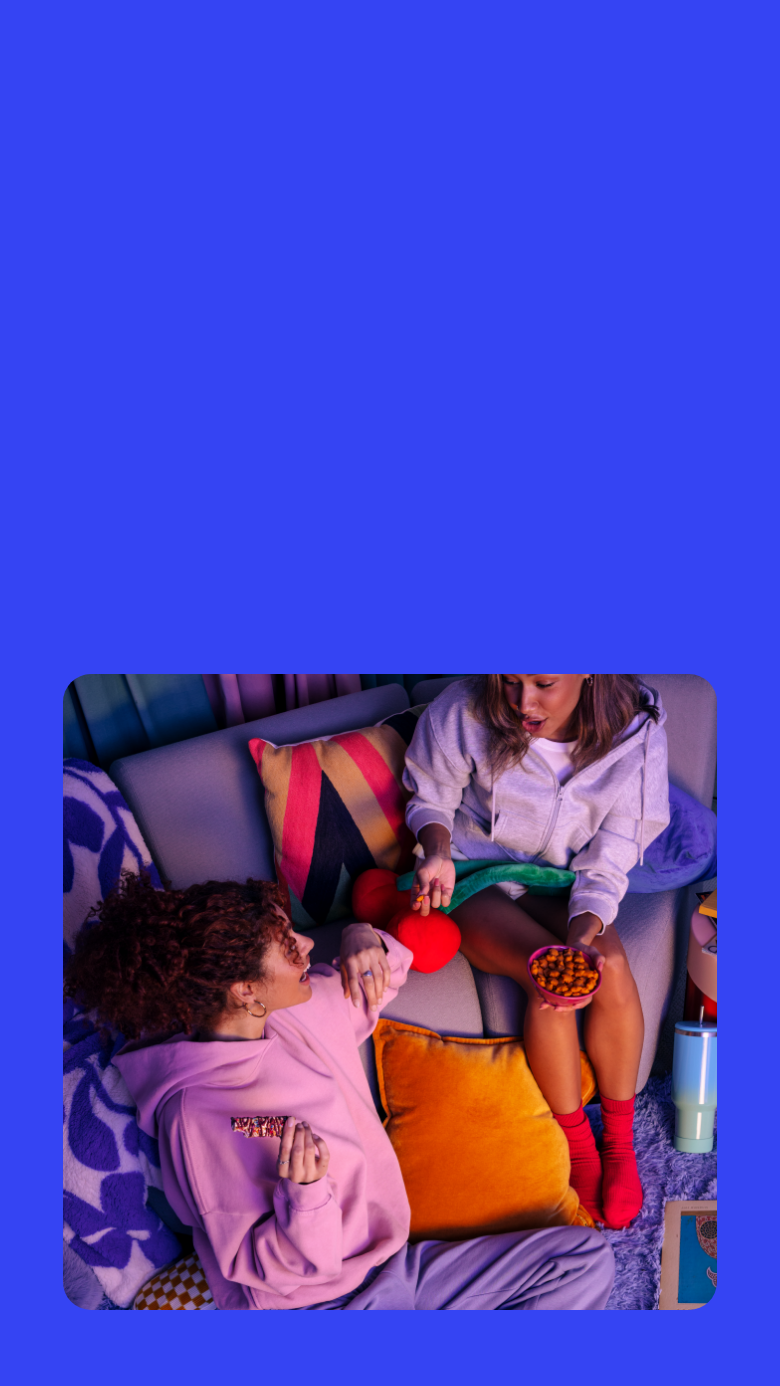 A woman in a pink hoodie lounges on a cozy couch, holding a colorful granola bar with a drizzle topping A woman in a pink hoodie lounges on a cozy couch, holding a colorful granola bar with a drizzle topping. She is engaged in conversation with another person, who is offering her a snack. The setting is warm and relaxed, with vibrant pillows and a plush cherry-shaped cushion adding to the cozy atmosphere.