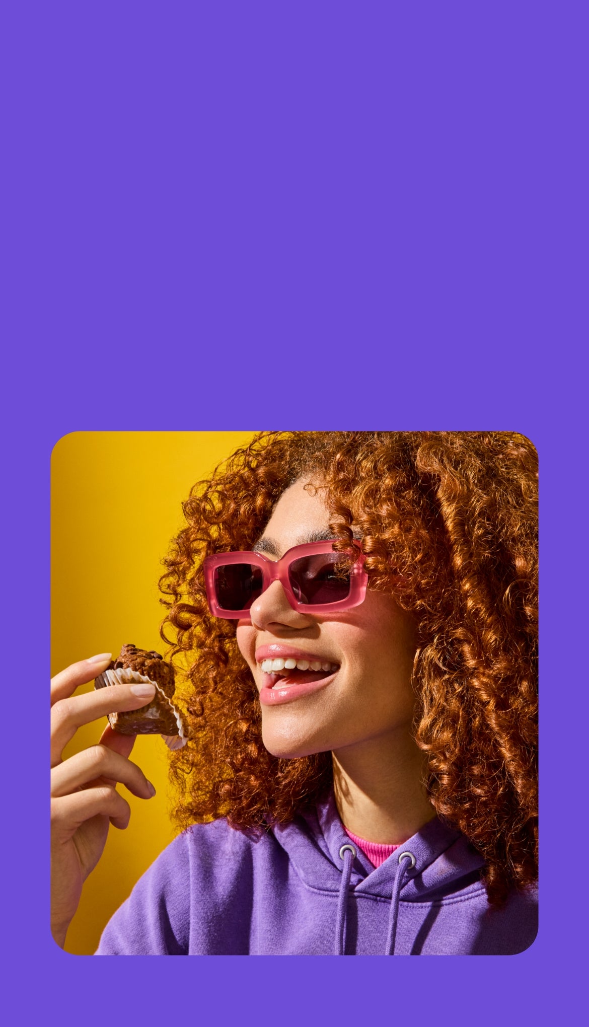 A young woman with voluminous curly red hair and pink sunglasses smiles as she holds a Mornings Oat Cup in a paper wrapper. She is wearing a purple hoodie and a pink turtleneck, with a bright yellow background adding vibrancy to the image. The scene exudes joy and indulgence.