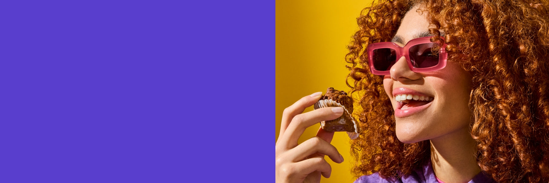 A young woman with voluminous curly red hair and pink sunglasses smiles as she holds a Mornings Oat Cup in a paper wrapper. She is wearing a purple hoodie and a pink turtleneck, with a bright yellow background adding vibrancy to the image. The scene exudes joy and indulgence.