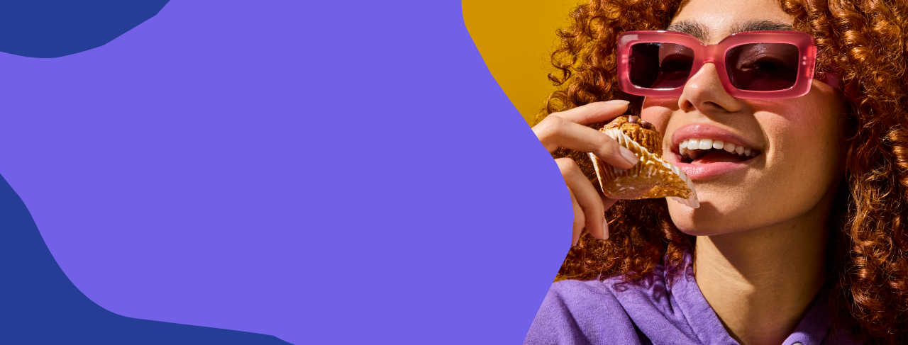 Woman eating a MadeGood Oat Cup while wearing pink sunglasses and a purple shirt against a blue and yellow abstract background.