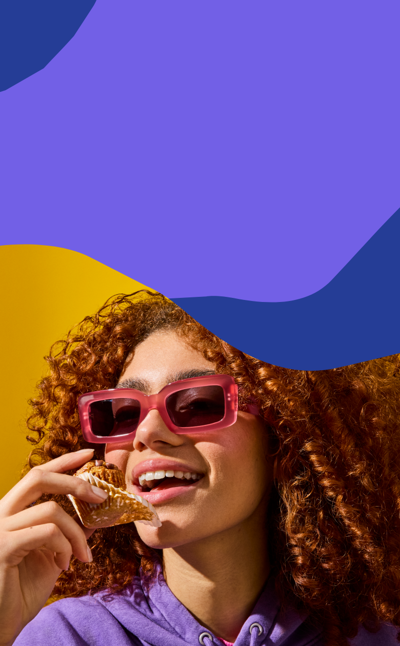 Woman eating a MadeGood Oat Cup while wearing pink sunglasses and a purple shirt against a blue and yellow abstract background.